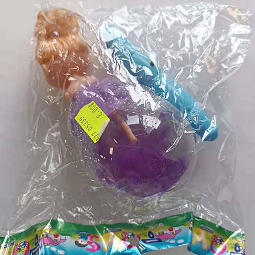 Păpușă de plastic într-o rochie textilă purpurie. Picioarele păpușii sunt plasate într-o minge de plastic transparentă cu pene. Un controler, inclusiv o baterie, este atașat la capul păpușii.